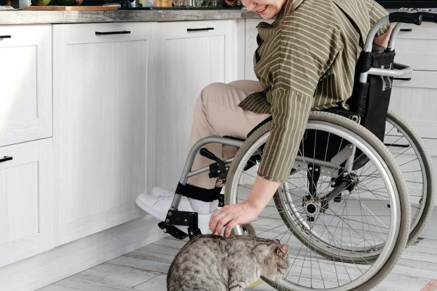 Pessoa em cadeira de rodas na cozinha inclinada para acariciar um gato