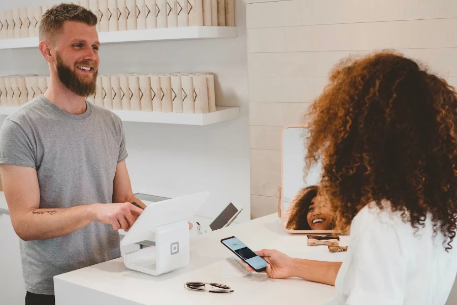 Smiling male shop assistant helps female customer checkout using phone