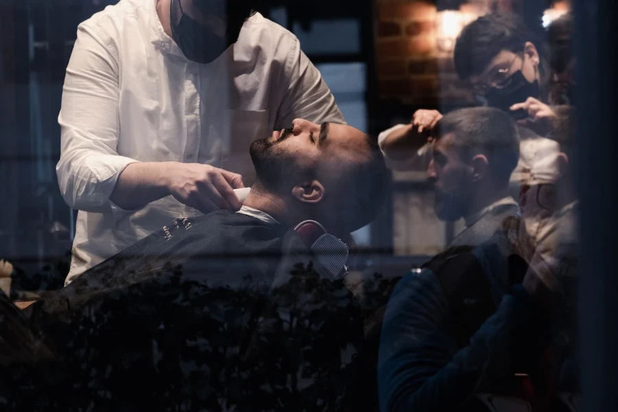 Deux hommes chez le coiffeur, l'un se faisant couper les cheveux, l'autre se faisant tailler la barbe