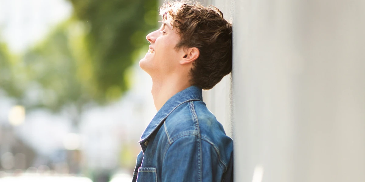 smiling young man in denim jacket leaning against a wall outdoors