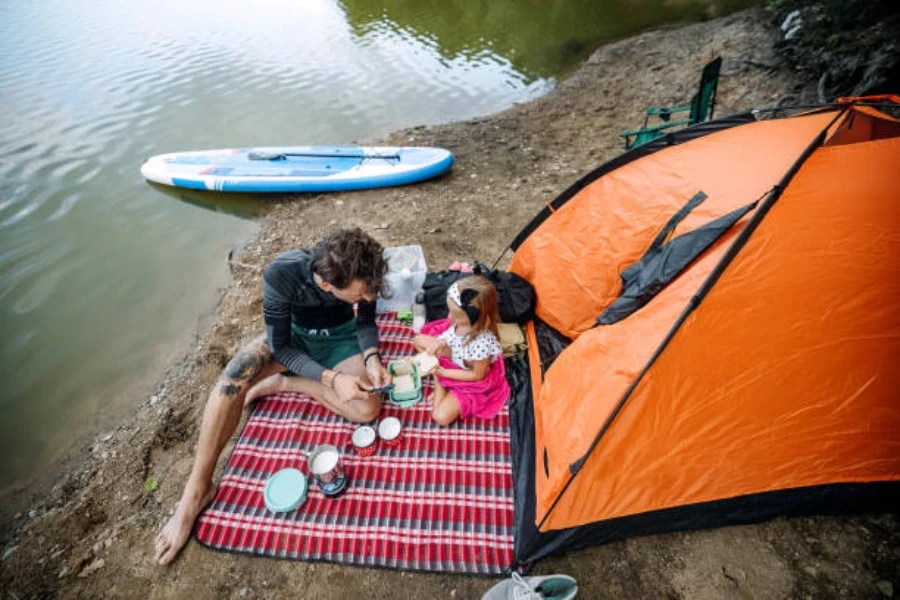 Man and daughter sitting on waterproof picnic blanket beside lake