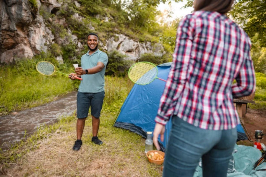 Man and woman playing badminton next to tent and stream