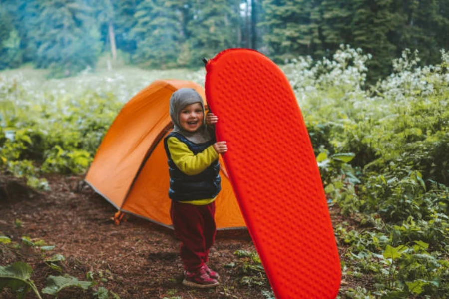 Criança pequena segurando uma esteira de acampamento vermelha na floresta