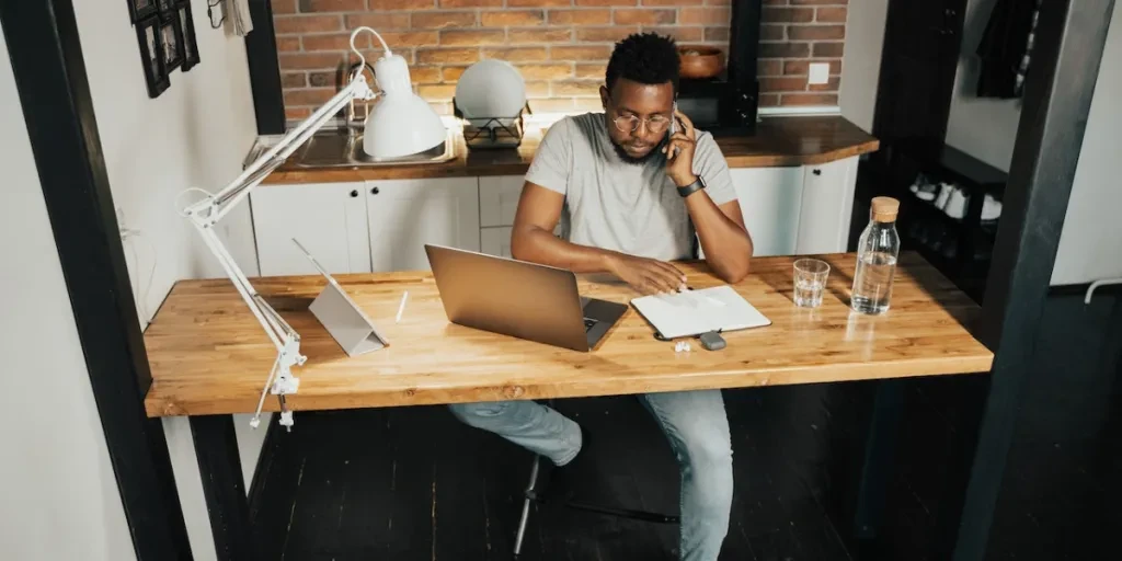 Pessoa falando ao telefone, sentada em uma mesa, trabalhando em frente a um laptop