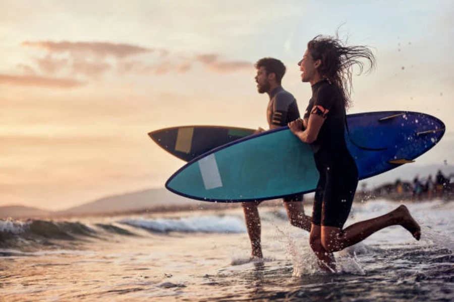 Hombre y mujer corriendo hacia el mar con tablas y trajes de neopreno