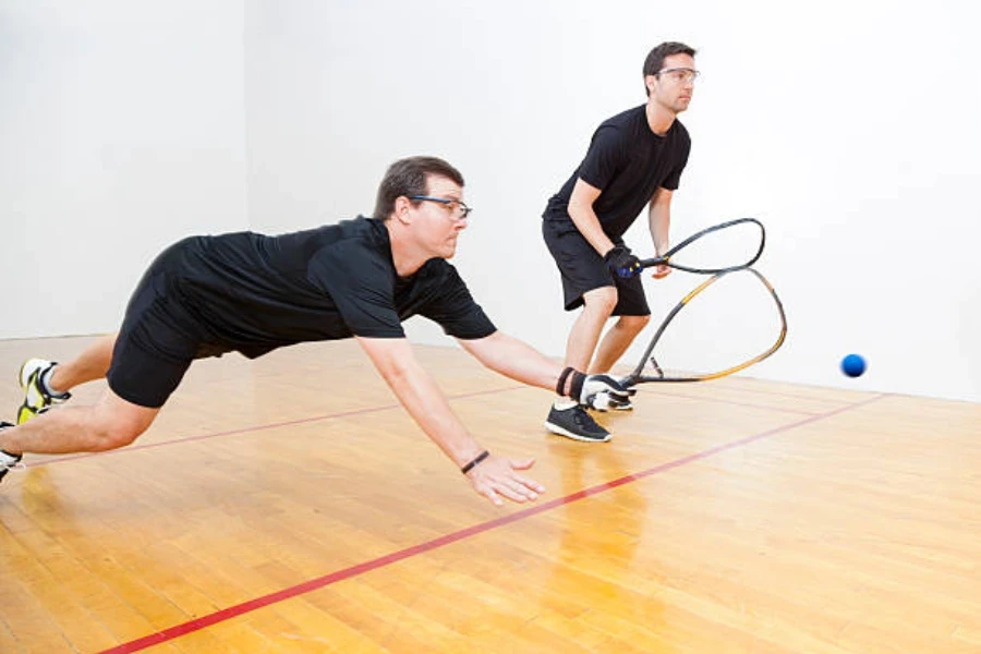 Dois homens usando óculos de squash e um deles mergulhando em busca de bola