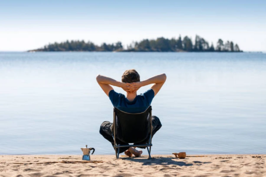 Mann sitzt im Campingstuhl am Strand und trinkt Kaffee