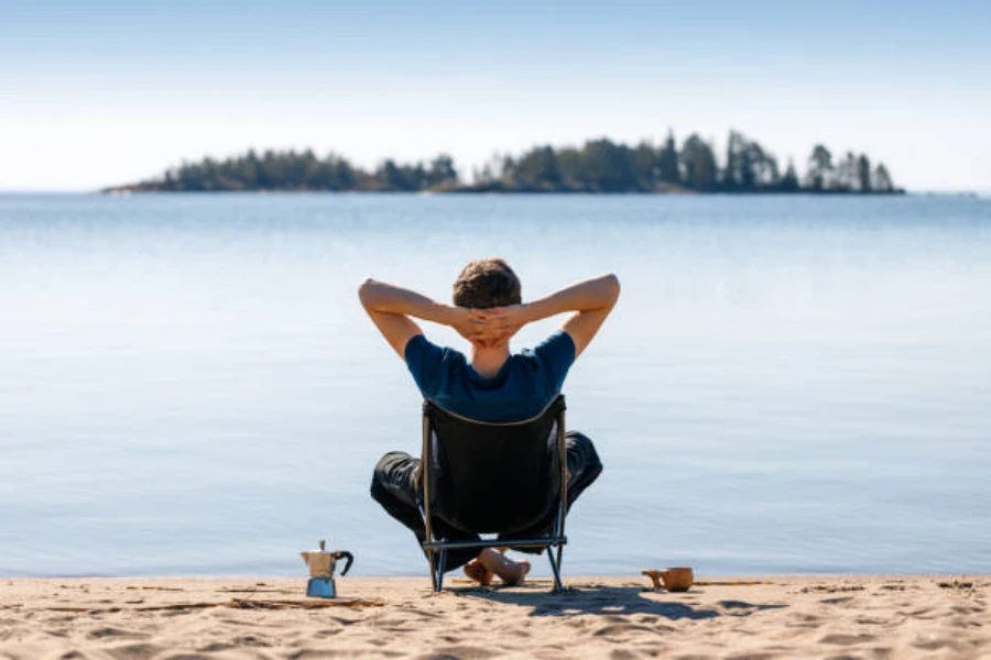 Pria yang duduk di kursi berkemah di pantai dengan kopi