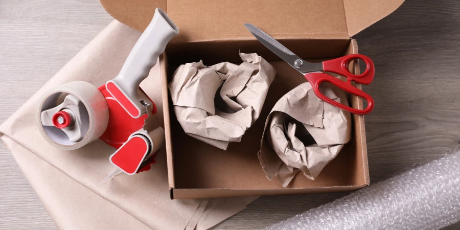 Offene Schachtel mit verpackten Gegenständen, Klebeband, Schere, Papier und Luftpolsterfolie auf einem Holztisch