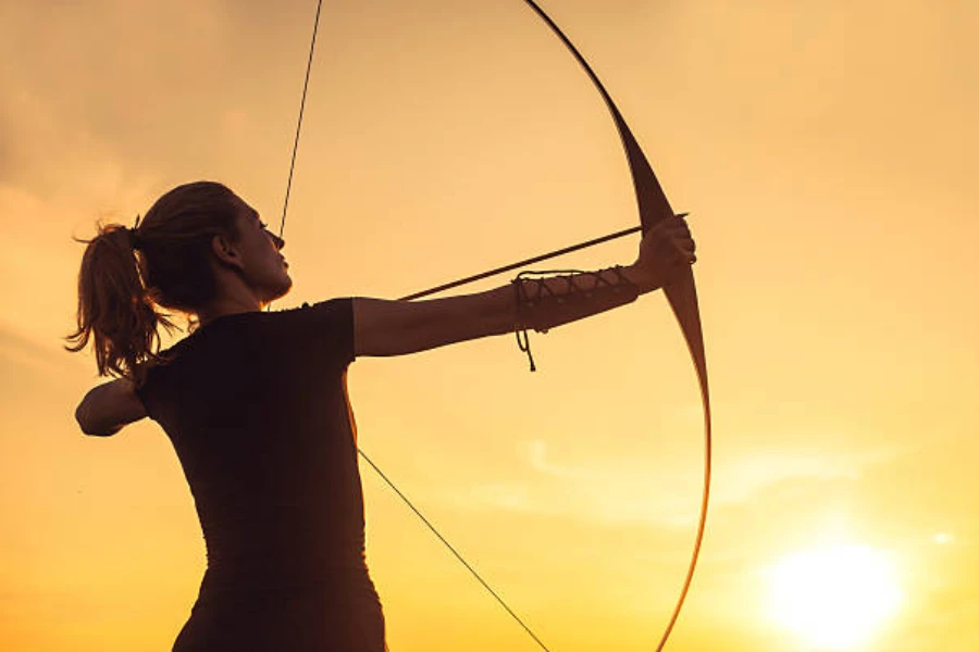 Woman using longbow to shoot arrow at sunset