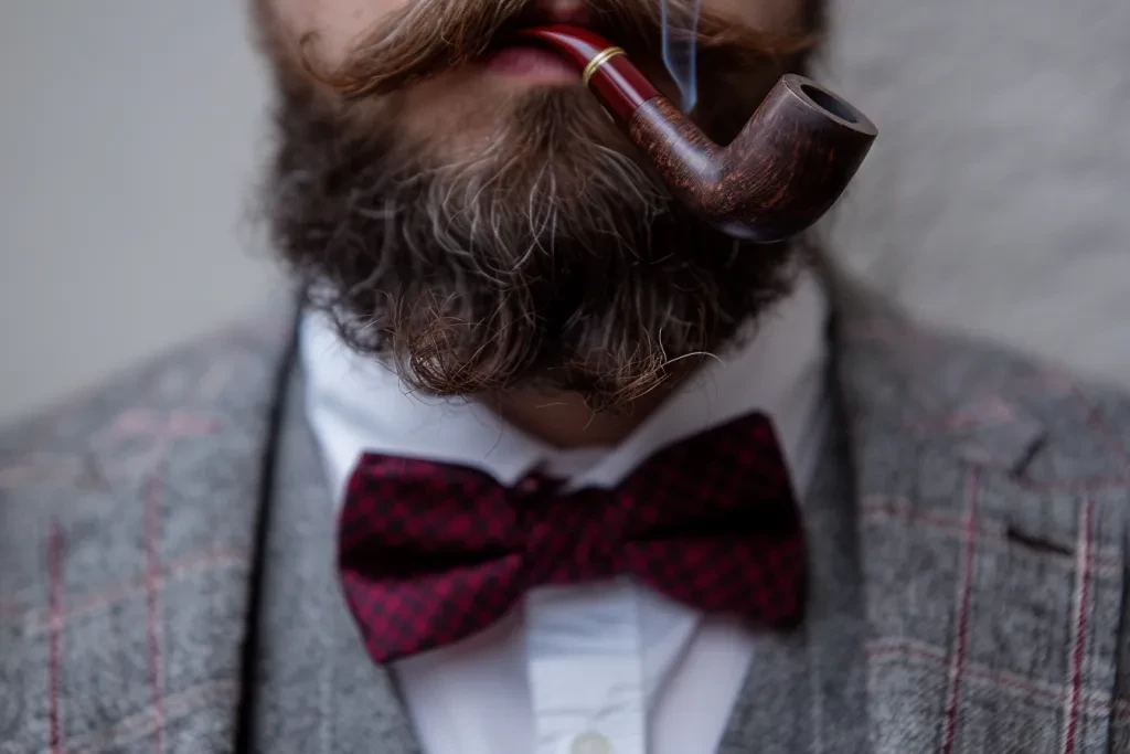 A man in grey suit and burgundy bow tie smoking pipe