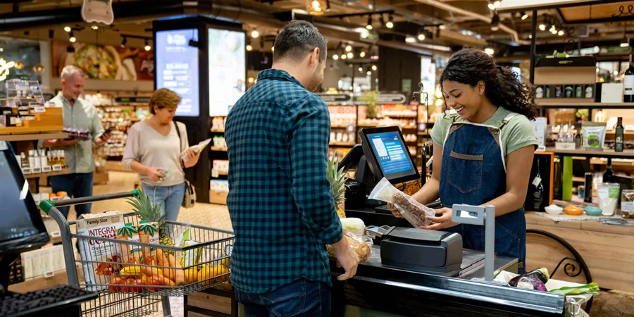 Feliz cajero afroamericano trabajando en el supermercado registrando productos y hablando con un cliente