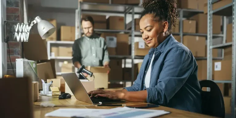 Lady types on a keyboard in her warehouse while a colleague moves boxes