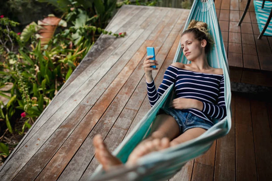 Mujer enviando mensajes de texto mientras está acostada en una hamaca plegable portátil azul