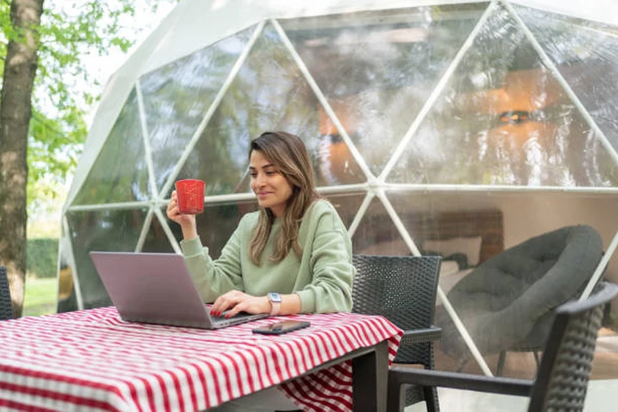 Wanita dengan cangkir merah duduk dengan laptop di lokasi glamping