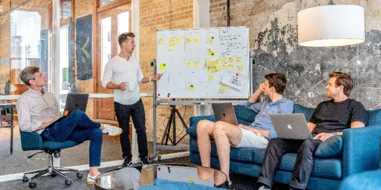 three men sitting while using laptops and watching man beside whiteboard