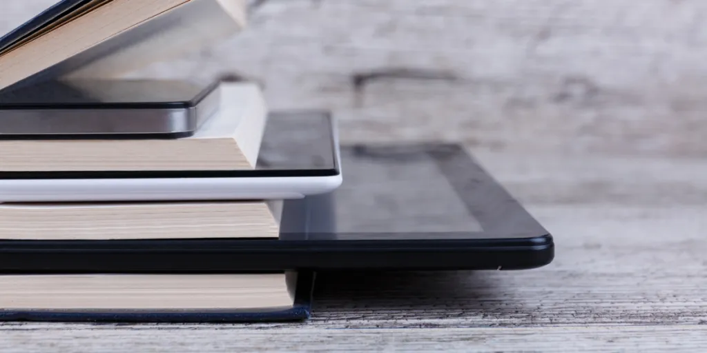 E-reader dan tablet di antara tumpukan buku
