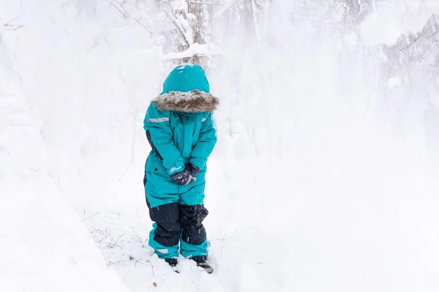 Un niño con un mono turquesa y capucha se encuentra en un bosque nevado y la nieve cae sobre él