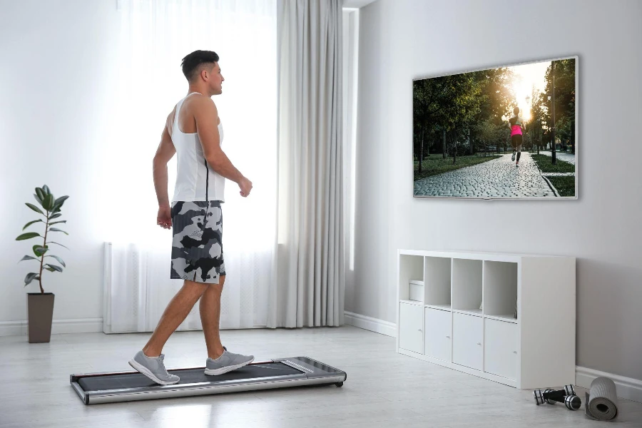 Спортивный мужчина тренируется на беговой дорожке и смотрит телевизор дома