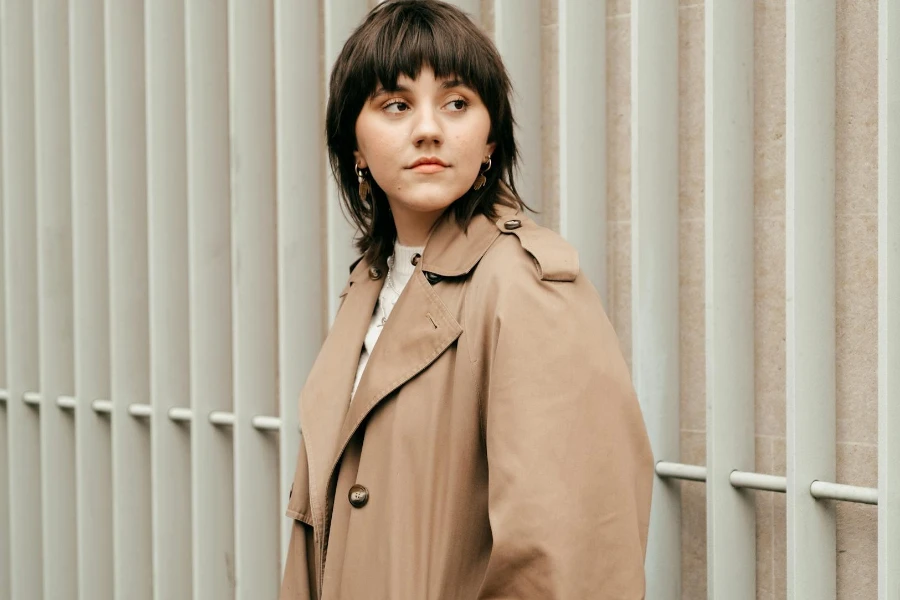 Calm young woman in trench coat looking away against wall in daytime