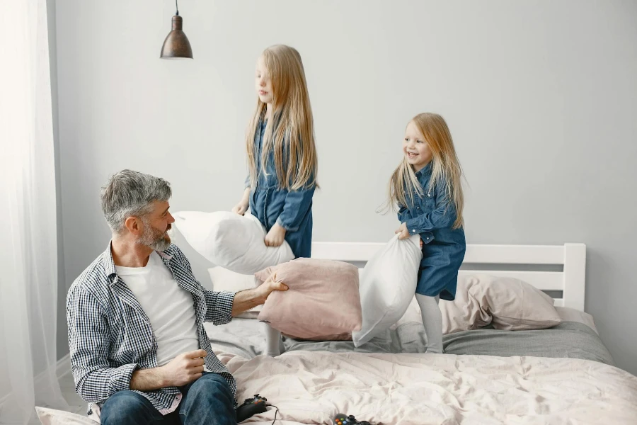 Girls in Denim Dress Standing on the Bed while Playing Pillows with their Grandfather 