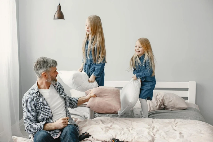 Girls in Denim Dress Standing on the Bed while Playing Pillows with their Grandfather 