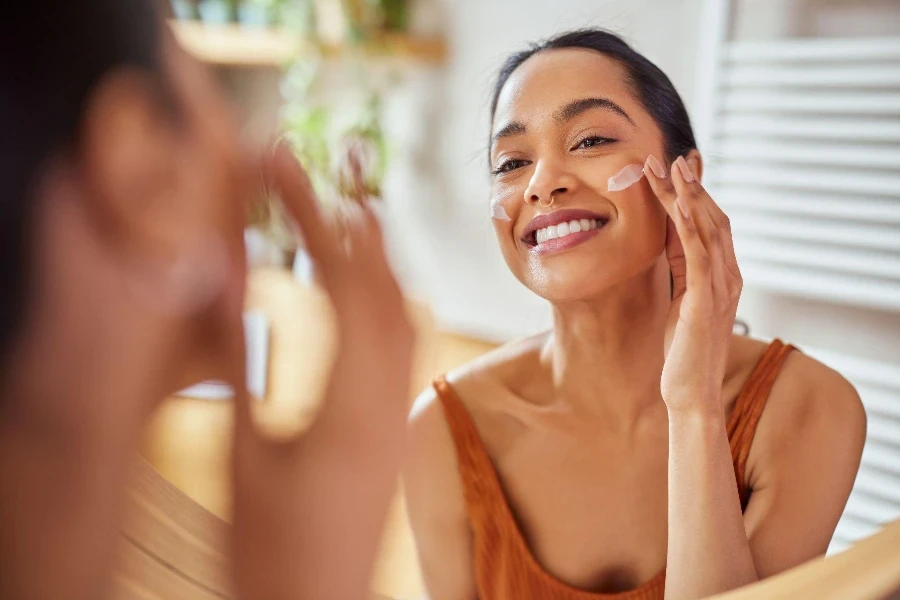 Uma mulher aplicando creme de mucina de caracol no rosto enquanto se olha no espelho