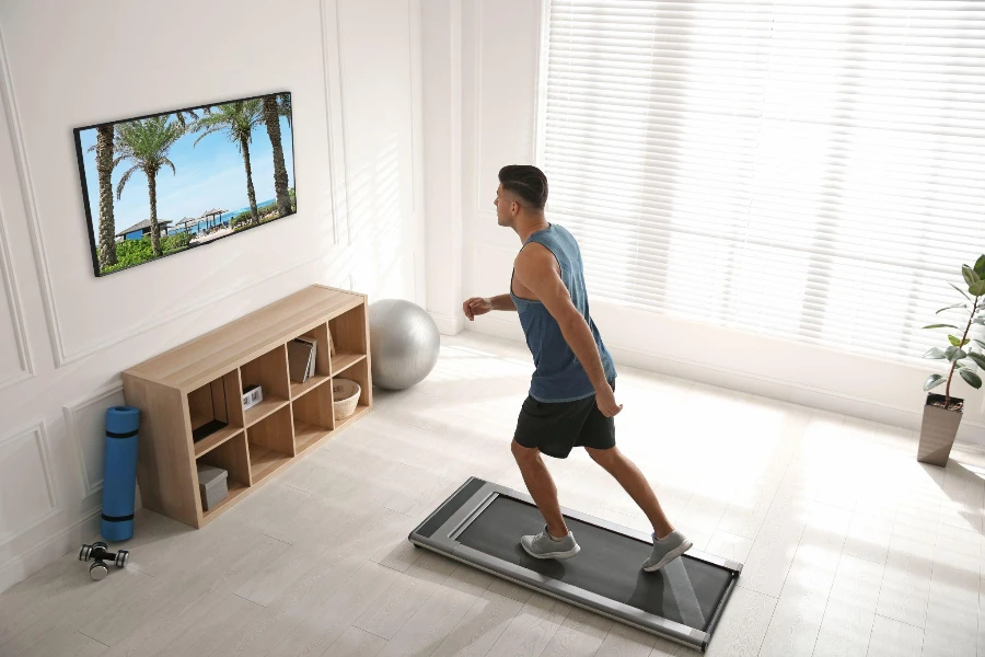 Hombre deportivo entrenando en una plataforma para caminar y viendo televisión en casa