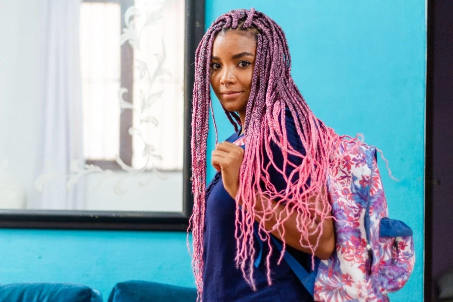 Young black woman with pigtails waving goodbye with colorful backpack leaving room with blue wall