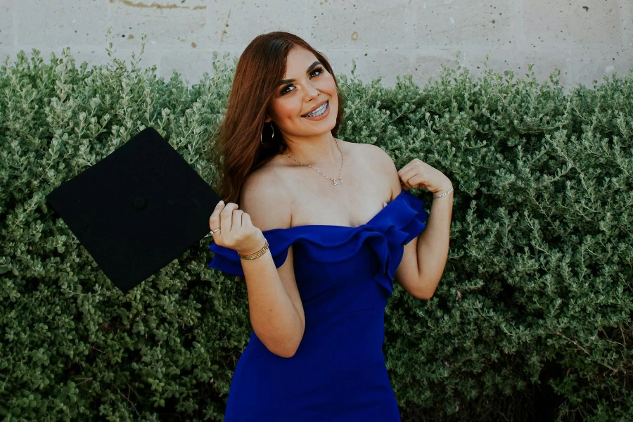 Cheerful female graduate holding academic cap next to brick wall