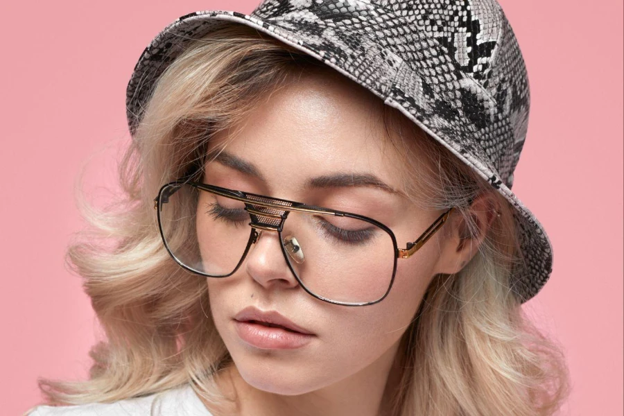 Modèle féminin blond sérieux dans un chapeau de seau tendance et des lunettes regardant vers le bas sur fond rose en studio