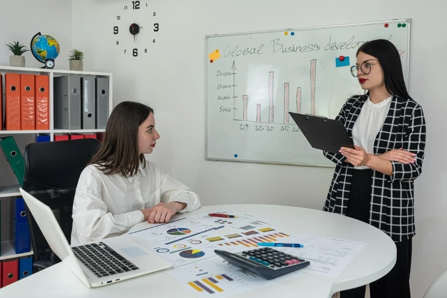 Two young casual female colleagues discussing near white board with new company business plan at office.