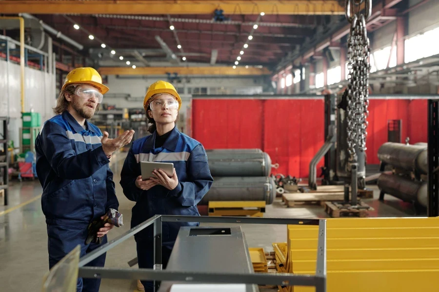 Bearded factory engineer in hardhat and safety goggles pointing at chains and explaining coworker how to use industrial crane