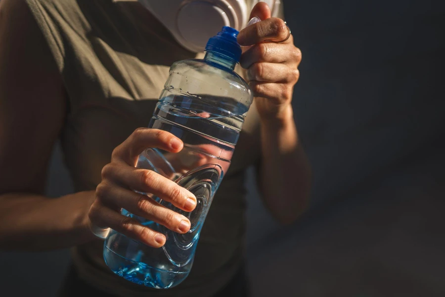 Nahaufnahme der Hände, Mittelteil einer Frau, offene Plastikflasche mit Wasser, während sie an sonnigen Tagen im Freien steht