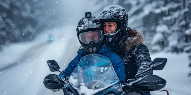 Un hombre con casco negro y traje de nieve azul está sentado en la parte trasera de la moto de nieve negra de su esposa.