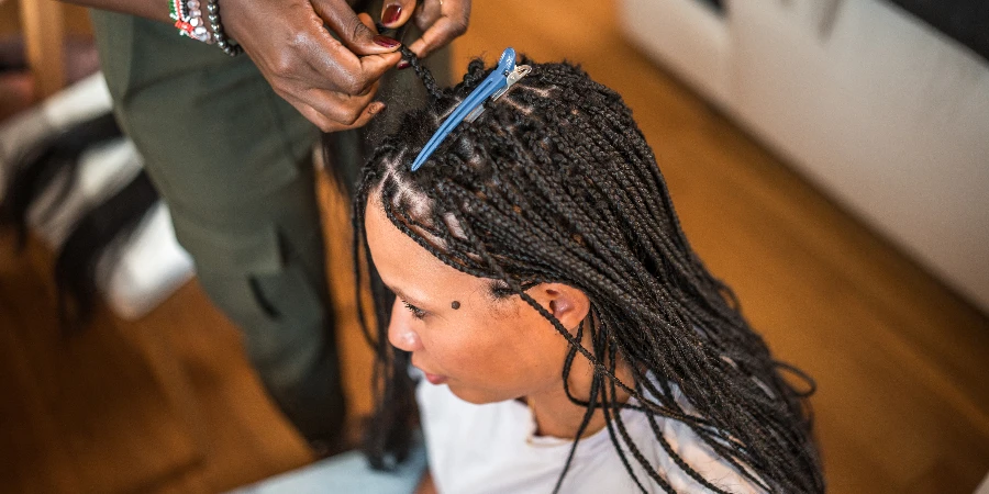 A professional mid-adult Black female hairstylist diligently works on braiding the hair of a Mixed race female client in a home setting