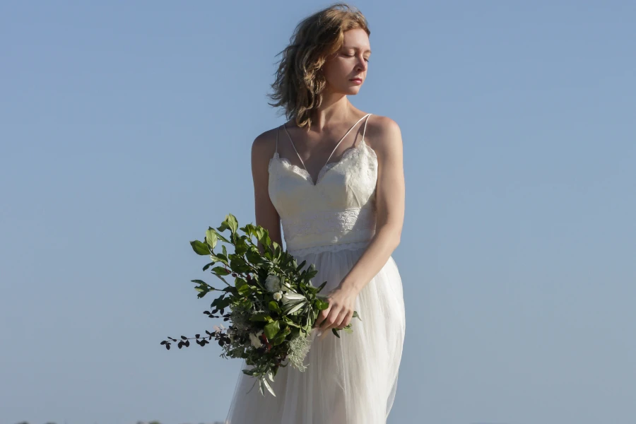 Bride in long white wedding dress standing on windy sea rock shore holding lush green wedding bouquet with boho twist