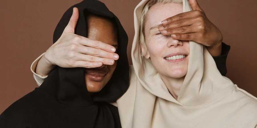 Content diverse female models in casual hoodies covering eyes of each other with hands while standing on brown background in studio