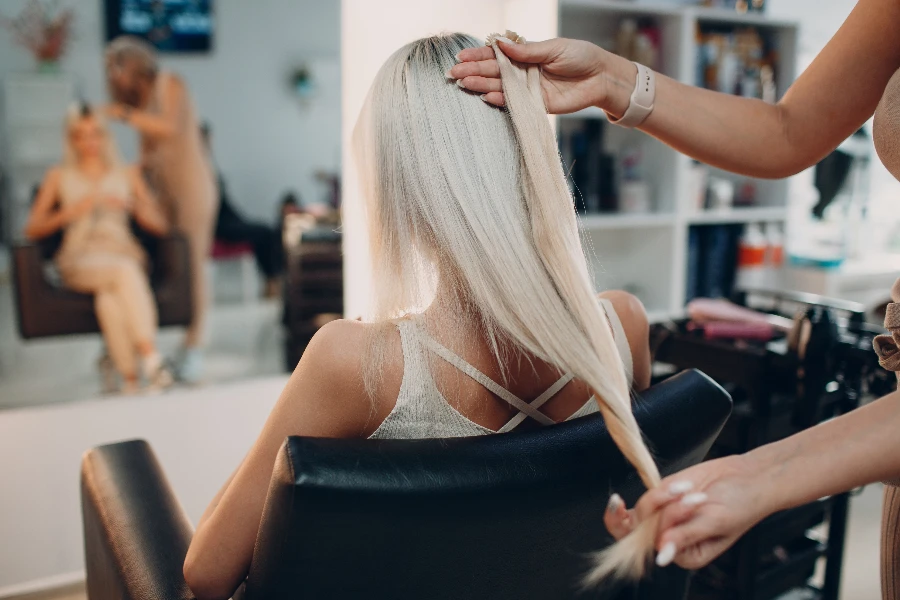 Hairdresser female making hair extensions to young woman with blonde hair in beauty salon