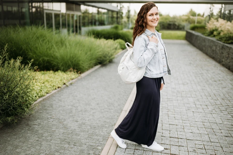 Glückliche hübsche junge Brünette Frau mit weißem Nylon-Rucksack in blauer Jeansjacke und blauem Maxirock im Freien