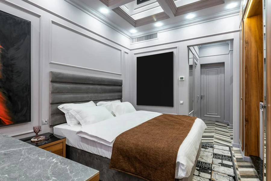 Modern hotel room with soft pillows and cover on bed between entrance door and table with glass