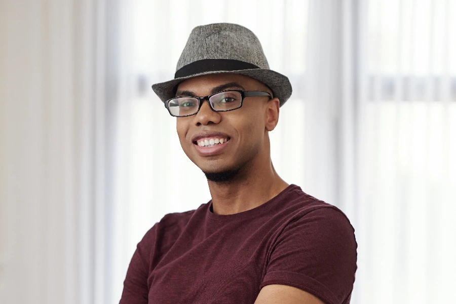 Portrait of happy young Black man in hat and glasses folding arms and smiling at camera