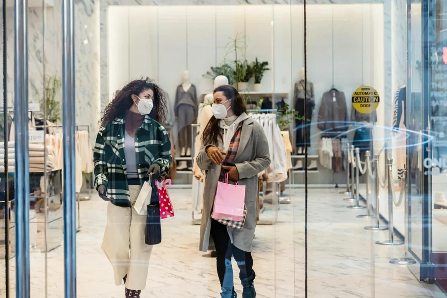 Through glass of female friends in protective masks carrying shopping bags while walking out of clothing store