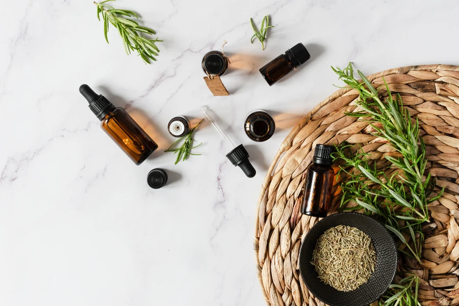 Bottles of rosemary essential oil and fresh rosemary branch on white marble background top view