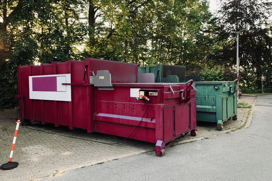 on an industrial site a green waste compactor and a red waste compactor stand side by side
