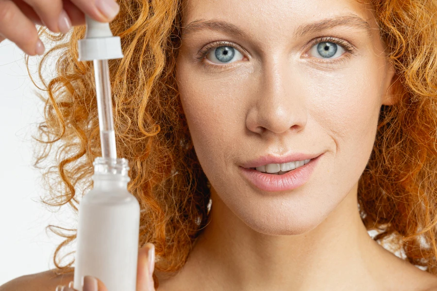 Close up portrait of a pleased red-headed lady with a bottle of cosmetic oil looking ahead