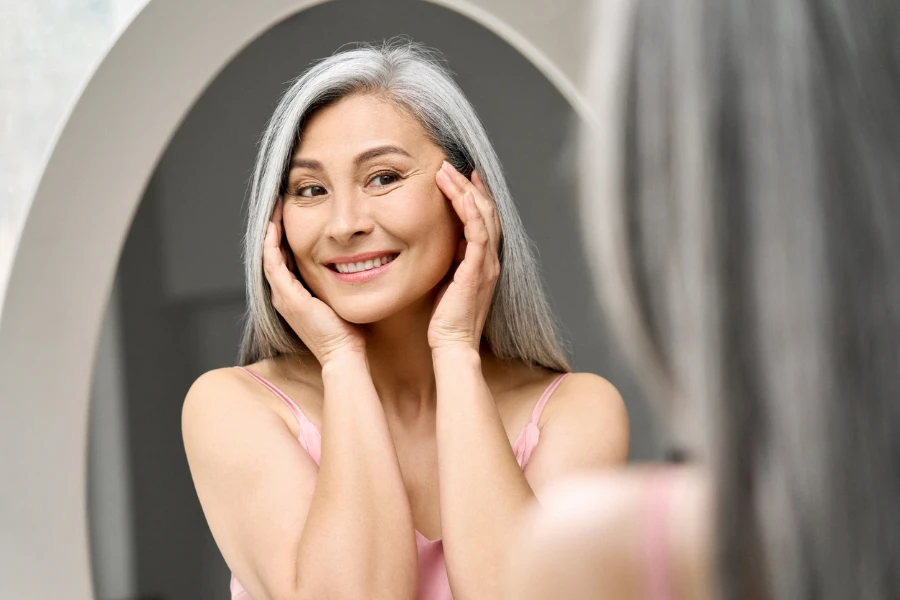 Happy middle 50 years aged asian woman with gray hair looking at mirror
