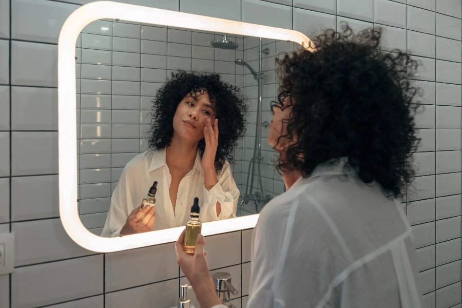 Young mixed race female gently applying face oil serum with fingers in modern bathroom