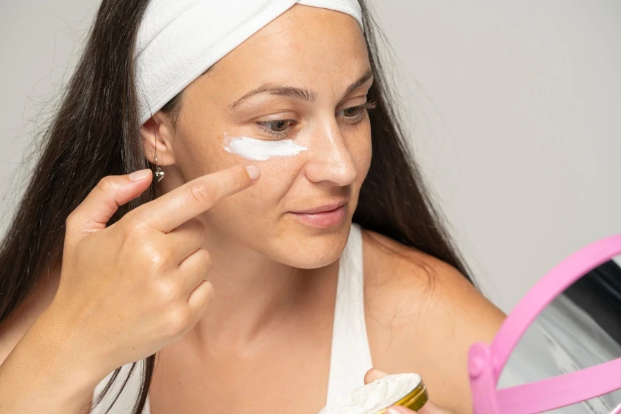 portrait of a young woman applying cosmetic product under her eyes on a gray background