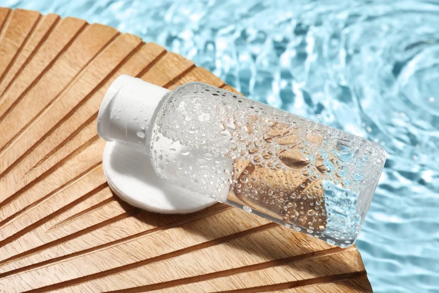 Wet bottle of micellar water and cotton pad on wooden board
